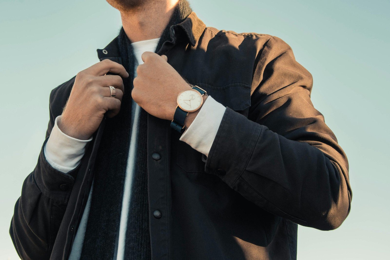 Man in a waxed cotton jacket and navy scarf adjusting his collar, wearing a rose-gold watch and a silver signet ring