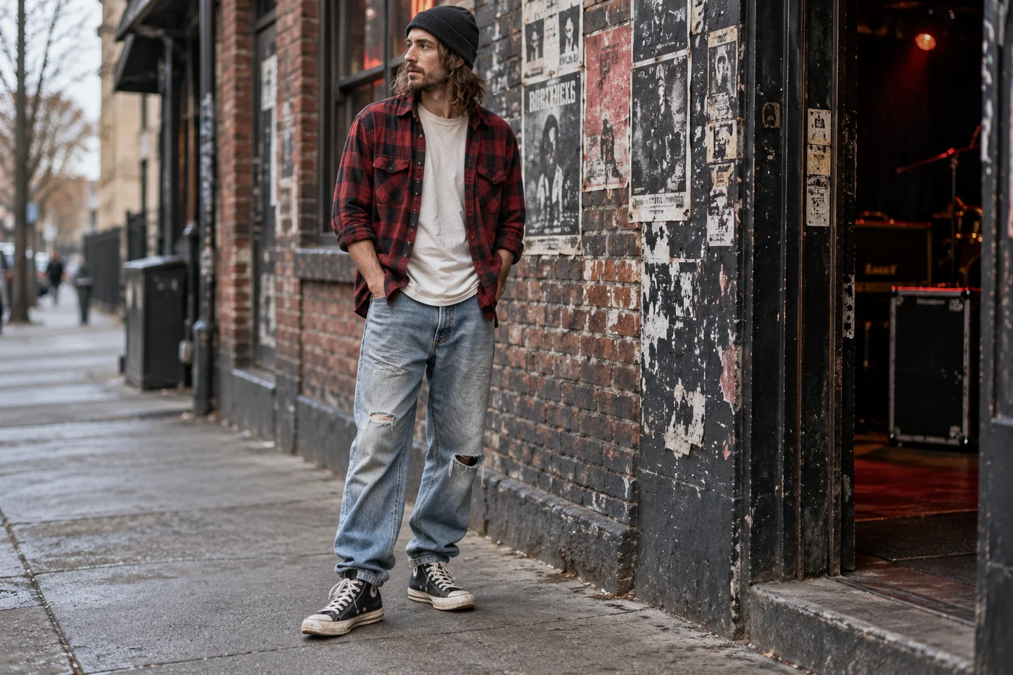 Man in red and black checked flannel, cream tee, loose faded jeans and black high-top trainers outside a music venue